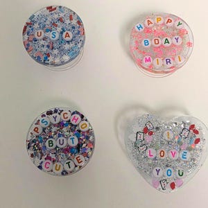 May include: Four decorative phone grips with glitter and text. One grip says "USA" with a patriotic theme. Another says "HAPPY BDAY MIRI". A third says "PSYCHO BUT CUTE". The last is heart-shaped and says "I LOVE YOU".