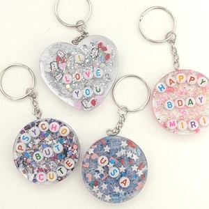 May include: A collection of handmade keychains with silver-colored key rings. The keychains are clear resin with glitter and small decorative elements. One is heart-shaped with "I LOVE YOU" spelled out. Others are round with phrases and names.