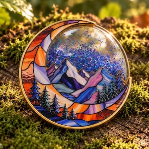 Op de afbeelding: Een ronde metalen speld met een ontwerp in glas-in-loodstijl van een maansikkel die een bergketen en dennenbomen omlijst. Het luchtgedeelte is gevuld met blauwe glitters, terwijl de maan en het landschap oranje, paarse en blauwe segmenten bevatten.