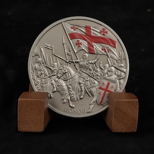 May include: A silver coin featuring knights on horseback, a flag with a red cross, and the word "HONOR". The coin is displayed on a wooden stand against a black backdrop. The flag has a red cross with four smaller crosses.