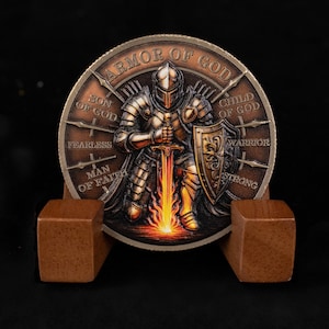 May include: A bronze-colored coin featuring a knight in full armor holding a sword with flames. The coin is inscribed with "ARMOR OF GOD" and other phrases. It rests on a wooden display stand against a black background.