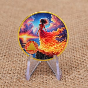 Puede incluir: Un medallón circular con borde dorado que presenta una ilustración colorida de una figura de pie sobre un acantilado al atardecer, con su vestido transformándose en llamas. Un emblema triangular dorado con el texto Unity, Service, Recovery.