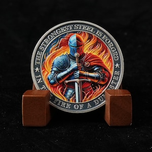 May include: A circular silver-toned coin featuring a knight in blue and red armour holding a sword, surrounded by orange and yellow flames. The border text reads, THE STRONGEST STEEL IS FORGED IN THE FIRE OF A DUNGEON MASTER.