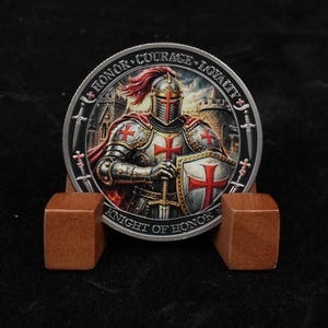 May include: A silver coin depicting a knight in armour, holding a sword and shield with a red cross. The coin is inscribed with "Honor, Courage, Loyalty" and "Knight of Honor". The coin is displayed on a wooden stand.