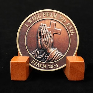 Könnte beinhalten: Eine bronzefarbene Münze mit dem Text "I WILL FEAR NO EVIL" und "PSALM 23:4", die ein Kreuz und betende Hände umgeben. Die Münze wird auf einem Holzständer mit zwei quadratischen Blöcken präsentiert.