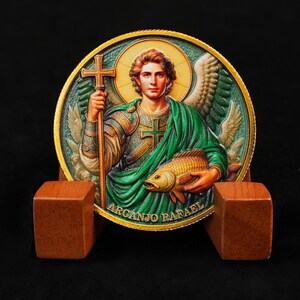May include: A detailed coin featuring Archangel Raphael holding a cross and a fish, with the text "Arcanjo Rafael". The coin has a gold rim and a green and gold background, and is displayed on a wooden stand.