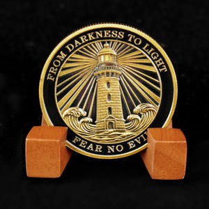 Peut inclure: Une pièce de monnaie dorée avec un phare, des vagues et les inscriptions "FROM DARKNESS TO LIGHT" et "FEAR NO EVIL". La pièce est présentée sur un présentoir en bois, sur fond noir.