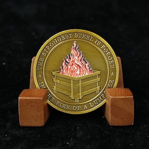 May include: A circular gold-toned metal coin featuring a raised design of a dumpster engulfed in red and orange flames. The text "THE STRONGEST STEEL IS FORGED IN THE FIRE OF A DUMPSTER" is embossed around the perimeter of the coin face.