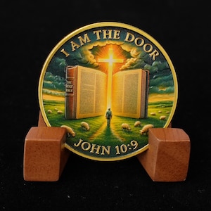 May include: A round, gold-colored coin with the text "I AM THE DOOR" and "JOHN 10:9". The coin features an open book, a cross, a person, and sheep in a field. The coin is displayed on a wooden stand.