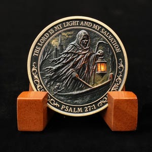 Può includere: Una moneta rotonda color bronzo raffigurante una figura incappucciata che tiene una lanterna in un paesaggio scuro. La moneta è incisa con "THE LORD IS MY LIGHT AND MY SALVATION" e "PSALM 27:1". È esposta su un supporto di legno.