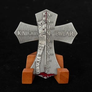 May include: A silver-colored Knights Templar cross with a detailed depiction of a knight and the words "KNIGHTS TEMPLAR" and "N.D.N.S.N.T.D.G." The cross is mounted on a wooden stand, with a red accent at the bottom.