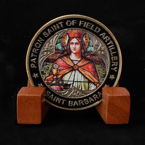 May include: A circular, gold-coloured coin depicting Saint Barbara, the patron saint of field artillery, with the text "PATRON SAINT OF FIELD ARTILLERY" and "SAINT BARBARA". The coin is displayed on a small, brown wooden stand against a black backdrop.