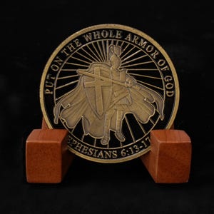 May include: A bronze-colored coin featuring a knight with a shield and sword, set on a wooden display stand. The coin has the text "PUT ON THE WHOLE ARMOR OF GOD" and "EPHESIANS 6:13-17".