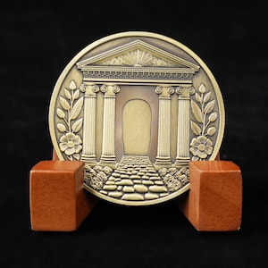 Peut inclure: Une pièce de monnaie de couleur bronze représentant une façade architecturale classique avec des colonnes et un chemin, ornée de motifs floraux. La pièce est posée sur un petit socle en bois brun-rouge, sur fond sombre.