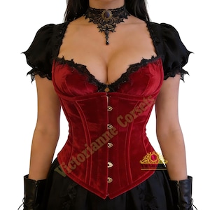 May include: A burgundy velvet corset with black lace trim and short black sleeves. The corset has a fitted silhouette with gold-colored clasps down the front. A black lace choker necklace with a pendant is also visible.