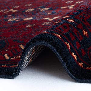 May include: Close-up of a rolled-up, intricately patterned rug. The rug features a deep red field with navy blue borders and accents. Small, light-colored geometric shapes are scattered across the red background, creating a detailed design. The rug's texture is visible.