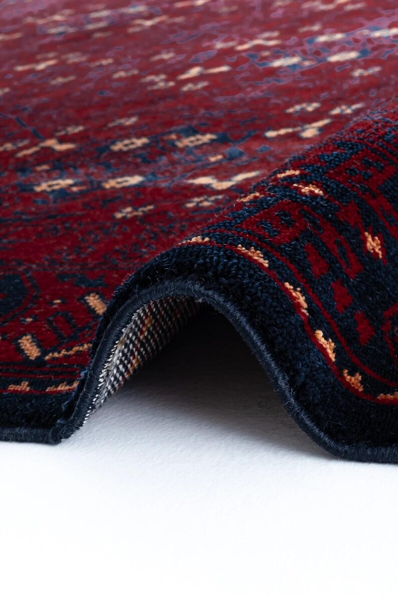 May include: Close-up of a rolled-up, intricately patterned rug. The rug features a deep red field with navy blue borders and accents. Small, light-colored geometric shapes are scattered across the red background, creating a detailed design. The rug's texture is visible.