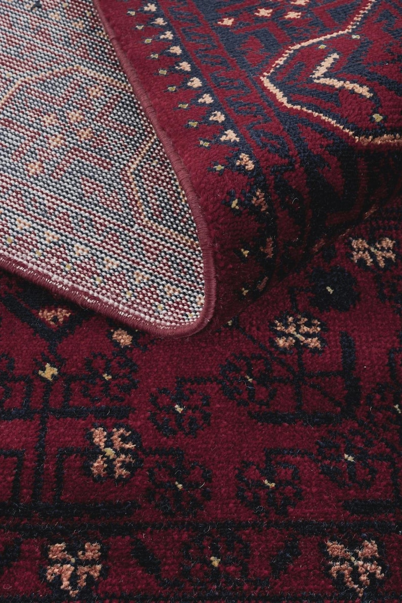 May include: Close-up of a burgundy-colored rug with an intricate pattern. The rug features a detailed design with dark blue and beige accents. The edges are neatly finished, showcasing the rug's quality and craftsmanship.