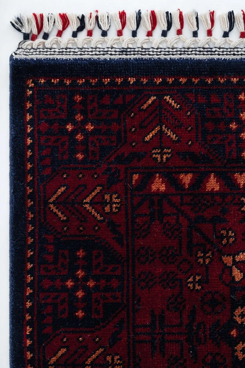 May include: Close-up of a section of a red and navy blue rug with an intricate geometric pattern. The rug has a dark navy blue border and is trimmed with white, red, and navy blue tassels. The design features repeating shapes and floral motifs.
