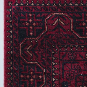 May include: Close-up of a section of a red rug with an intricate geometric pattern in black and gold. The rug has a fringe of white, red, and black tassels along the edge. The design features repeating shapes and borders.