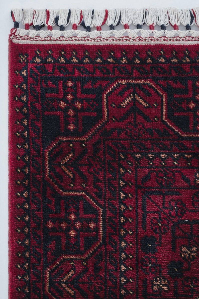 May include: Close-up of a section of a red rug with an intricate geometric pattern in black and gold. The rug has a fringe of white, red, and black tassels along the edge. The design features repeating shapes and borders.