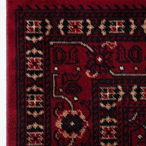 May include: Close-up of a red rug with a detailed geometric pattern in black and tan. The rug has a border with repeating shapes and a fringe along the top edge. The rug's design is intricate and traditional.