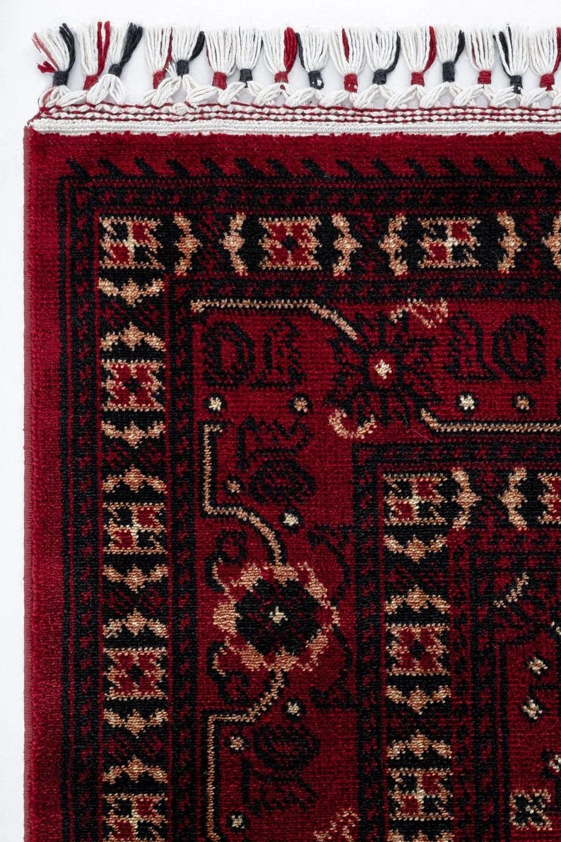 May include: Close-up of a red rug with a detailed geometric pattern in black and tan. The rug has a border with repeating shapes and a fringe along the top edge. The rug's design is intricate and traditional.