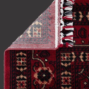 May include: A section of a red oriental rug with a detailed floral pattern in black and gold. The rug's edge features a white border with tassels in red, black, and white. The underside of the rug is visible, showing a geometric design.