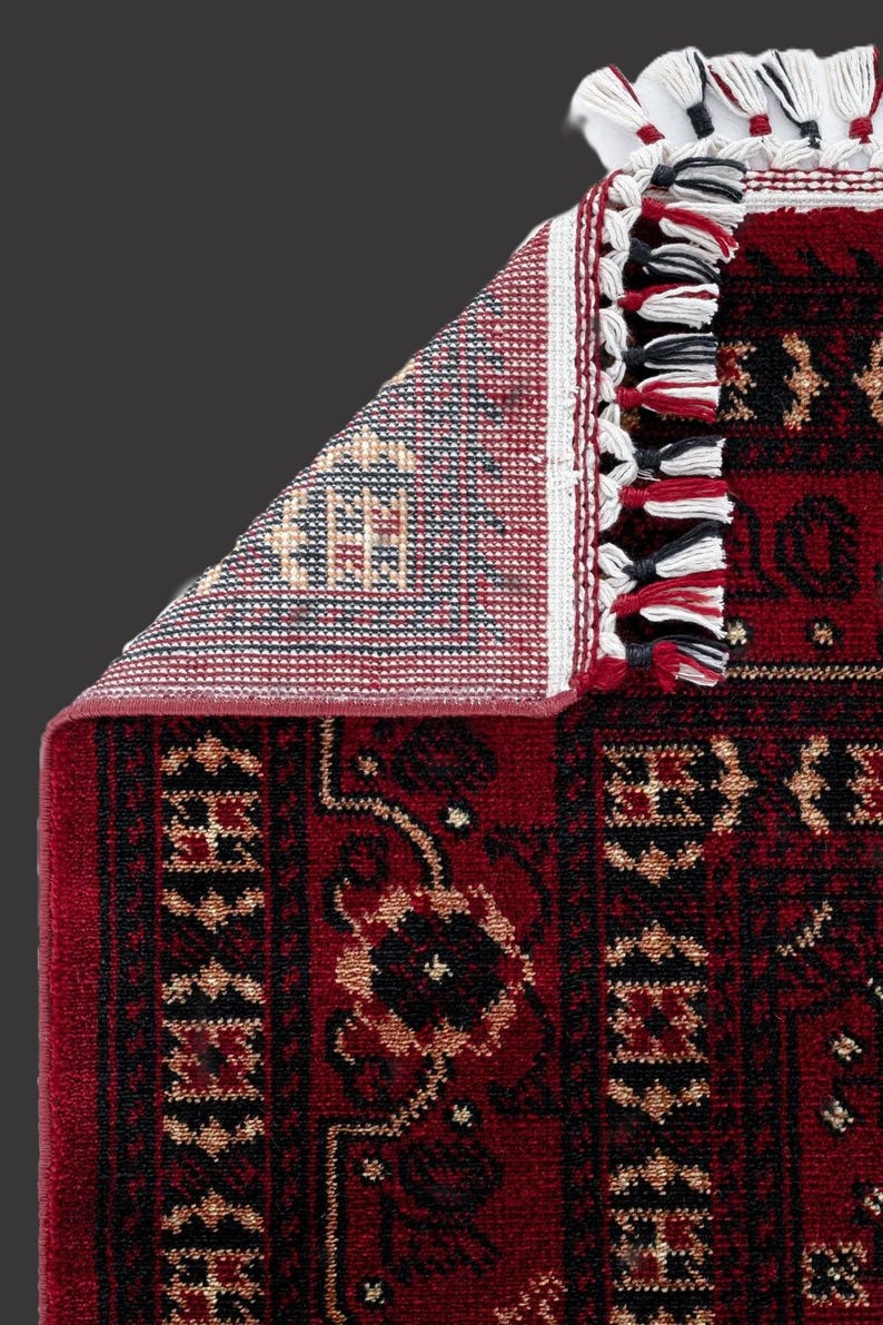 May include: A section of a red oriental rug with a detailed floral pattern in black and gold. The rug's edge features a white border with tassels in red, black, and white. The underside of the rug is visible, showing a geometric design.