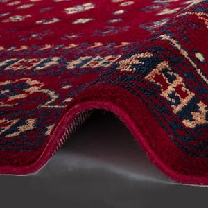 May include: Close-up of a rolled-up red rug with an intricate pattern. The rug features a deep red background with a repeating design in navy blue and beige. The edges are neatly finished, showcasing the rug's texture.