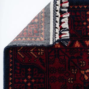 May include: A section of a red and navy blue rug with a detailed geometric pattern. The rug has a navy blue border and white, red, and navy blue tassels. The underside of the rug is visible, showing a striped pattern in shades of orange, red, and black.