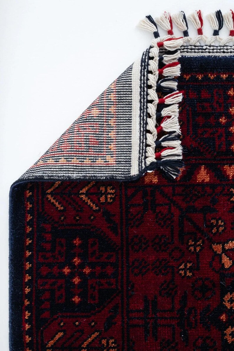 May include: A section of a red and navy blue rug with a detailed geometric pattern. The rug has a navy blue border and white, red, and navy blue tassels. The underside of the rug is visible, showing a striped pattern in shades of orange, red, and black.