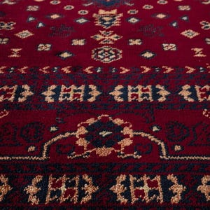 May include: Close-up of a red rug with an intricate geometric pattern. The design features navy blue and beige accents, creating a detailed and textured surface. The rug's pattern includes diamond and floral motifs, suitable for home decor.