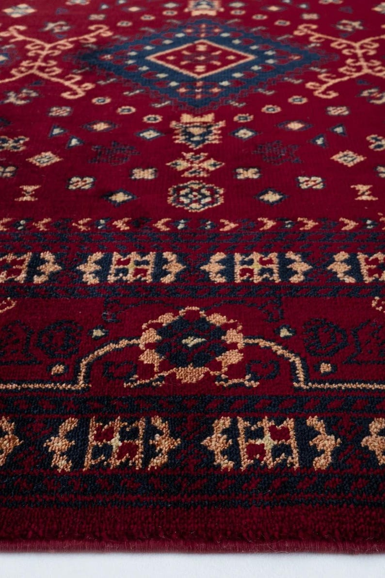 May include: Close-up of a red rug with an intricate geometric pattern. The design features navy blue and beige accents, creating a detailed and textured surface. The rug's pattern includes diamond and floral motifs, suitable for home decor.