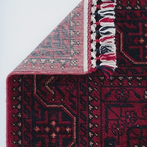 May include: A section of a red and black patterned rug with a geometric design. The rug has a detailed border with white, red, and navy blue tassels. The corner of the rug is folded back, revealing the underside with a patterned design.