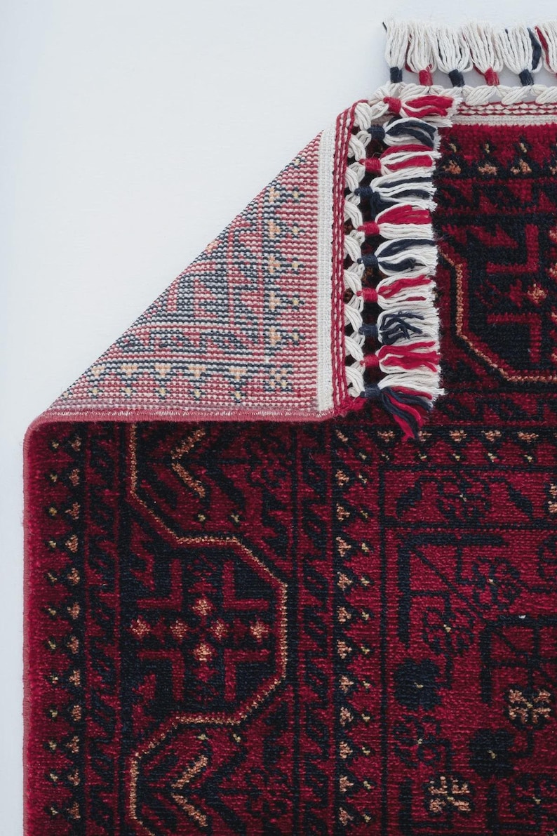 May include: A section of a red and black patterned rug with a geometric design. The rug has a detailed border with white, red, and navy blue tassels. The corner of the rug is folded back, revealing the underside with a patterned design.