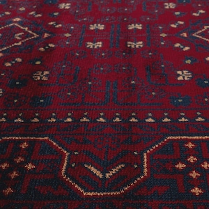 May include: Close-up of a red rug with a detailed geometric pattern. The rug features a deep red background with navy blue and tan accents. The design includes repeating floral and geometric shapes.