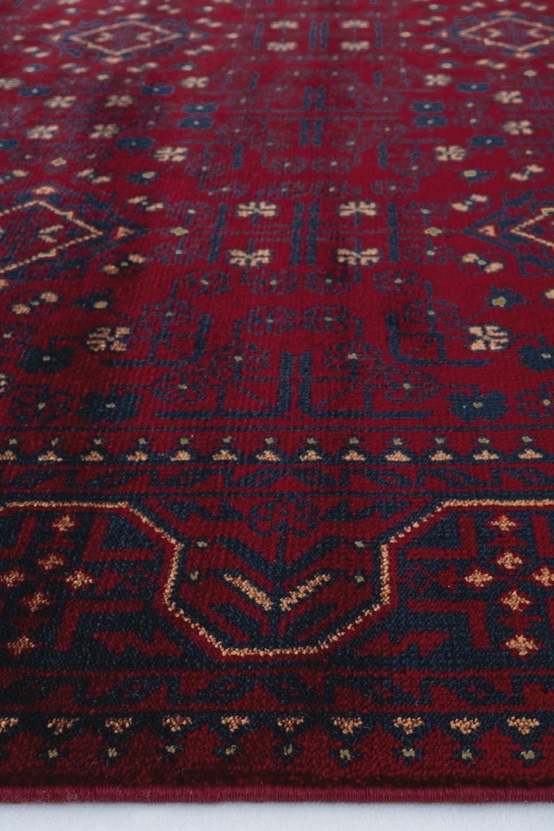May include: Close-up of a red rug with a detailed geometric pattern. The rug features a deep red background with navy blue and tan accents. The design includes repeating floral and geometric shapes.