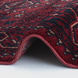 May include: Close-up of a rolled-up red rug with a detailed pattern. The rug features a deep red background with intricate designs in navy blue and beige. The edges are neatly finished with a red border, showcasing the rug's texture and craftsmanship.