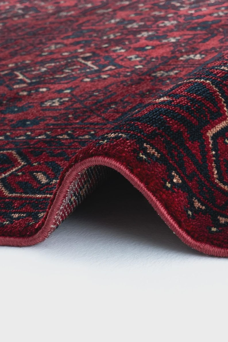 May include: Close-up of a rolled-up red rug with a detailed pattern. The rug features a deep red background with intricate designs in navy blue and beige. The edges are neatly finished with a red border, showcasing the rug's texture and craftsmanship.