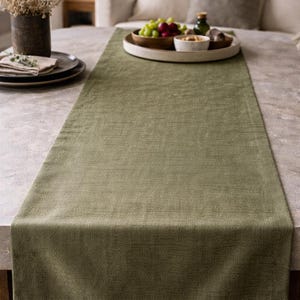 Op de afbeelding: Een saliegroene tafelloper van textiel. De loper is rechthoekig en ligt op een lichtgekleurde tafel. De tafel is gedekt met een houten dienblad met druiven, een kommetje en een fles.