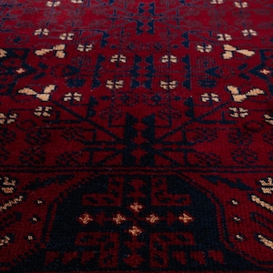 May include: Close-up of a red and navy blue patterned rug. The rug features a repeating geometric design with a dark red background and navy blue and tan accents. The rug's texture is visible.
