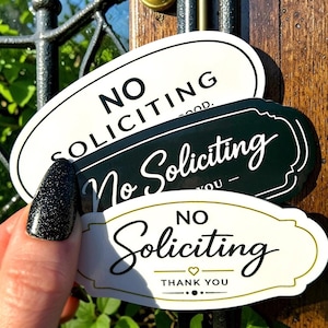 Könnte beinhalten: Drei ovale und rechteckige Schilder mit der Aufschrift „No Soliciting“ in Weiß, Dunkelgrün und Creme. Die Schilder haben elegante Schriftarten und Ränder. Auf einem Schild steht unten „Thank You“. Die Schilder werden vor einer Holztür gehalten.