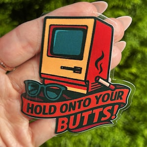 May include: A sticker featuring a retro computer design in yellow and red, with sunglasses and a lit cigarette. The sticker has the text "HOLD ONTO YOUR BUTTS!" in red on a banner.
