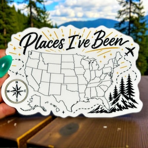 Peut inclure: Un autocollant blanc avec le texte "Places I've Been" en écriture cursive, représentant une carte des États-Unis. Le design comprend une boussole, un avion et une chaîne de montagnes avec des arbres.