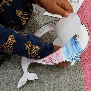 K&ouml;nnte beinhalten: Ein Papierbastelprojekt mit einem ausgeschnittenen Wal in Blau und Rosa. Der Wal hat eine bewegliche Klappe am Kopf. Ein gedrucktes Anleitungsblatt mit dem Titel JONAH AND THE WHALE CRAFT und dem Text Color, cut, fold and glue ist zu sehen.