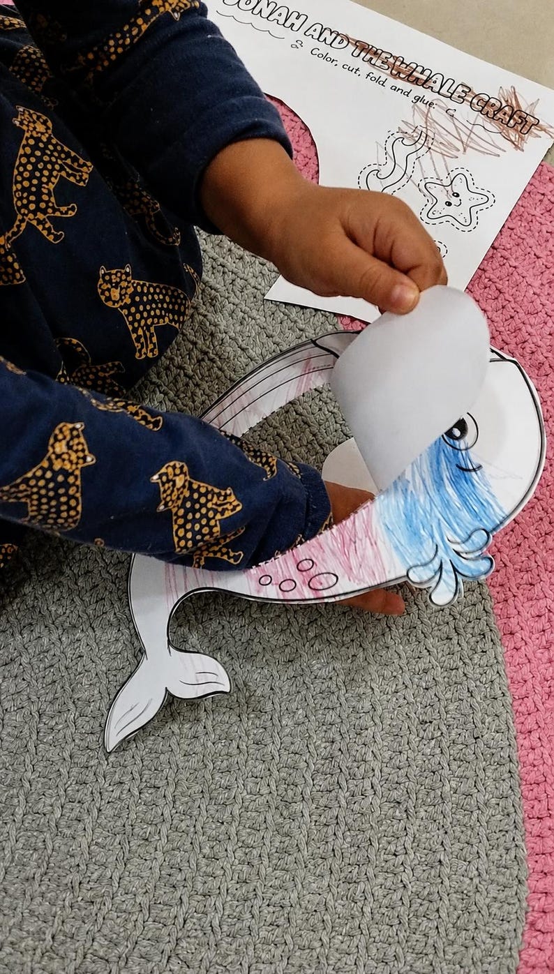 K&ouml;nnte beinhalten: Ein Papierbastelprojekt mit einem ausgeschnittenen Wal in Blau und Rosa. Der Wal hat eine bewegliche Klappe am Kopf. Ein gedrucktes Anleitungsblatt mit dem Titel JONAH AND THE WHALE CRAFT und dem Text Color, cut, fold and glue ist zu sehen.