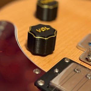 Pode incluir: Close-up de um botão de volume de guitarra preto com acabamento dourado e a palavra "VOL" em letras douradas. O botão está no corpo de madeira de uma guitarra com acabamento vermelho. Outro botão é visível no fundo.