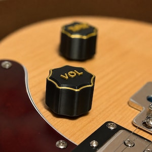 Puede incluir: Dos perillas de control negras estriadas con borde y letras doradas sobre un cuerpo de guitarra de madera clara. La perilla frontal presenta el texto VOL en dorado, mientras que la perilla trasera tiene texto dorado parcialmente visible.