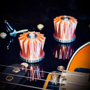 Pode incluir: Close-up do corpo de uma guitarra, mostrando dois botões de controle laranja e prateados. Os botões têm um design único, com nervuras e refletem-se na superfície preta brilhante. As cordas da guitarra e uma parte do corpo de madeira também são visíveis.